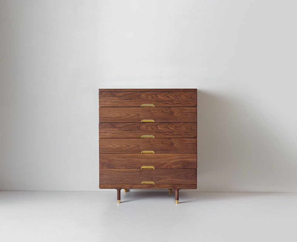 A wooden chest of drawers with brass handles against a light wall, viewed full-frontal.
