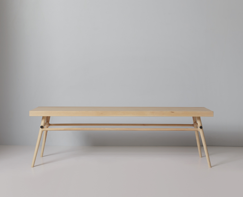 The same wooden bench shown in a room with light wooden floor; a long low wooden table with books rests on the bench.