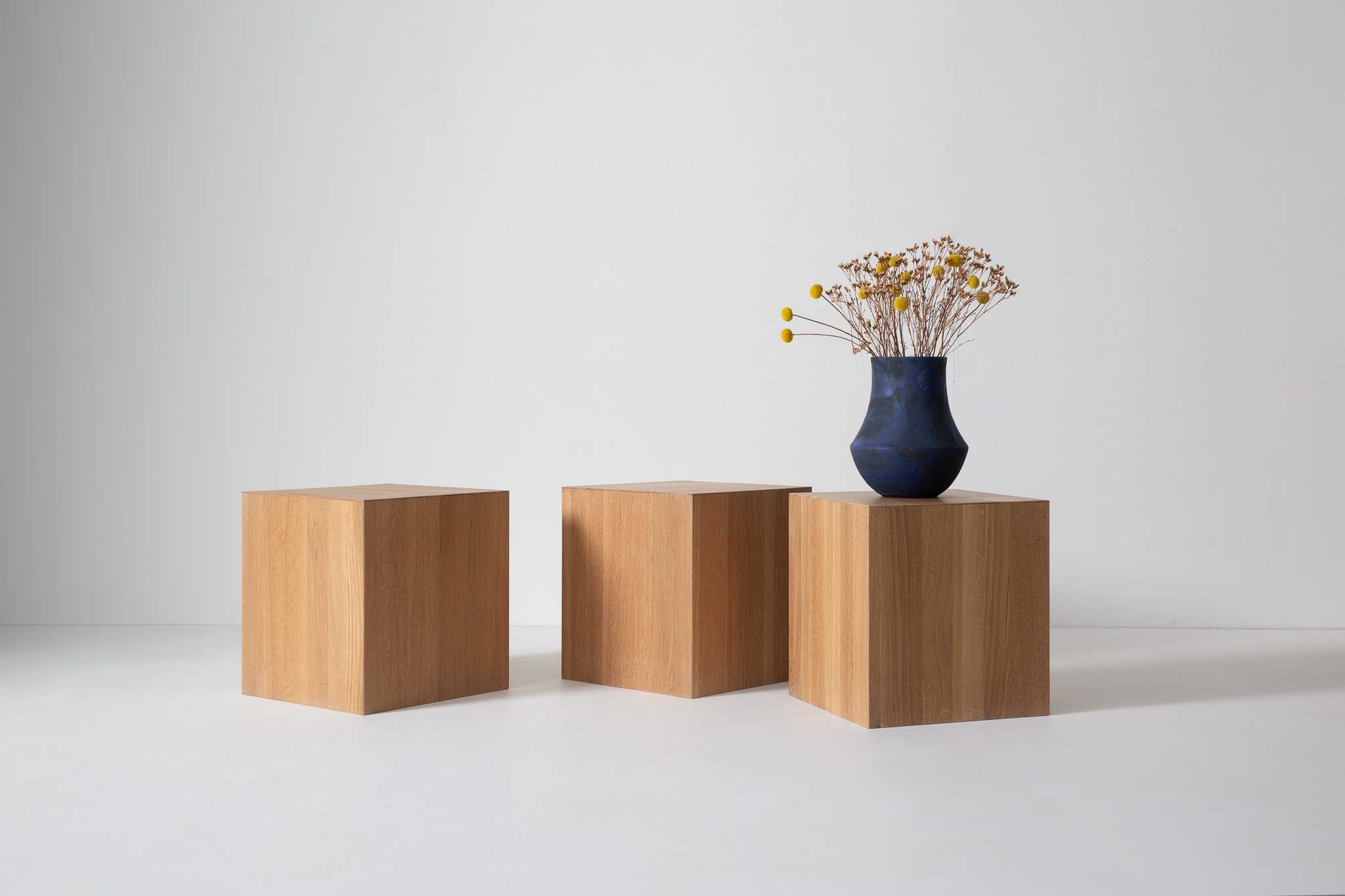 A blue vase with dried yellow flowers placed on a wooden cube pedestal, against a white wall.