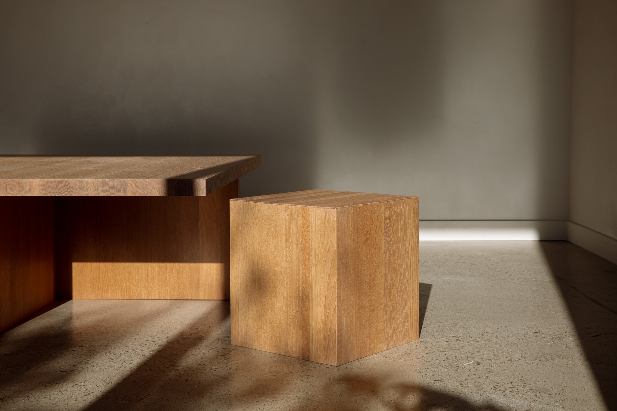 A single wooden cube sits on a concrete floor with strong light and shadow across the surface.